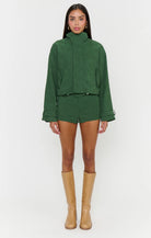 A model stands against a light gray studio background wearing a matching dark green cropped jacket and shorts set, tan knee-high boots, and gold hoop earrings.