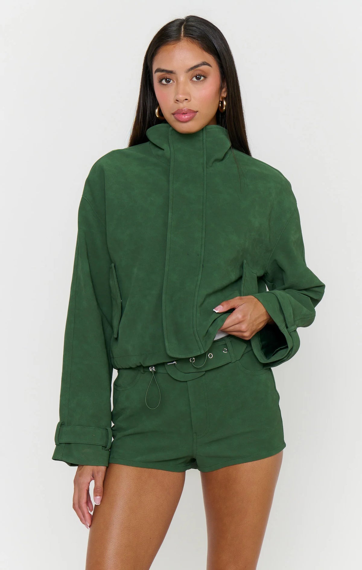 A woman modeling a dark green cropped hooded jacket with a high collar and matching green shorts against a light gray studio background.