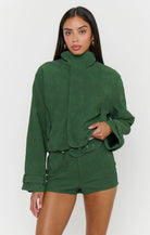 A woman modeling a dark green cropped hooded jacket with a high collar and matching green shorts against a light gray studio background.