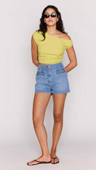 Woman wearing a yellow off-shoulder top and blue denim shorts on a white background