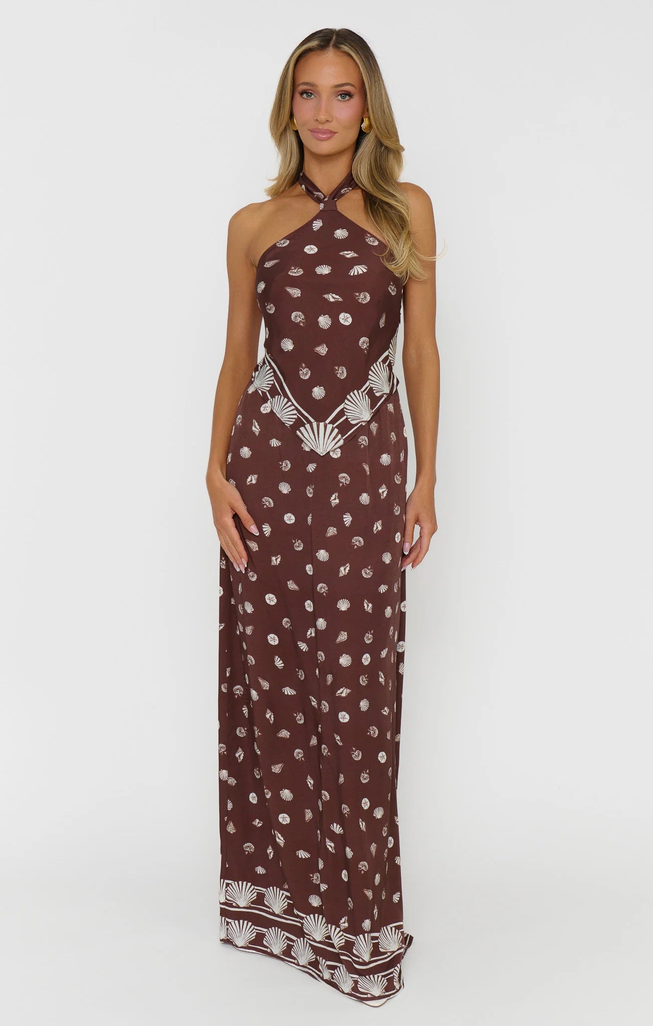A woman modeling a halter-neck maxi dress in a dark brown color with a white bohemian-style print and patterned hem, set against a plain light gray background.