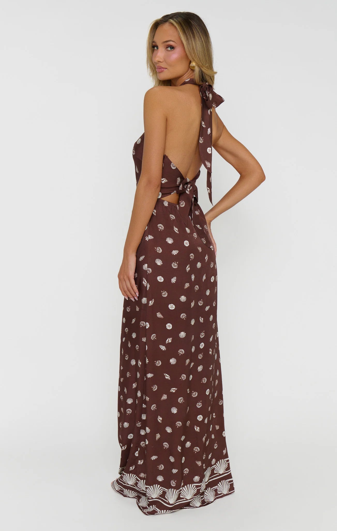 A woman modeling a brown halter-neck maxi dress with a white floral print and tie-back straps, shown from the back against a plain white studio background.