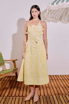 Woman wearing a yellow and white striped dress standing on a wooden floor with a chair and striped canopy in the background.