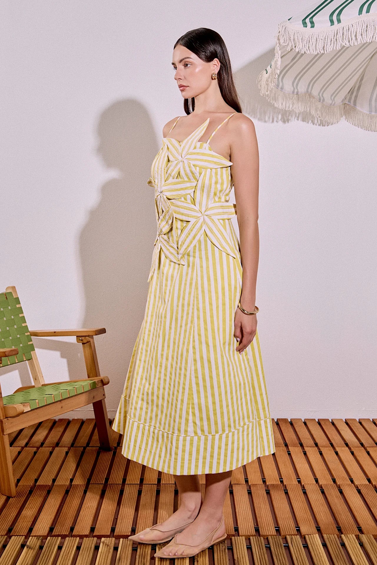 Woman wearing a yellow and white striped dress standing on a wooden floor with a chair and table in the background.