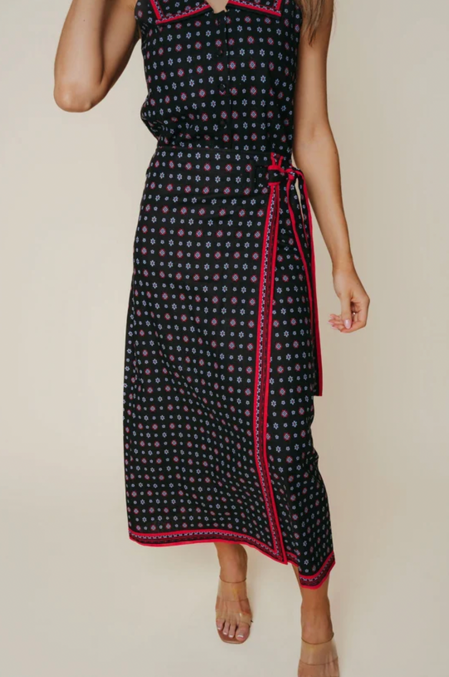 A sleeveless midi dress with a black base, white and pink floral pattern, and red trim, shown on a model against a plain beige background.