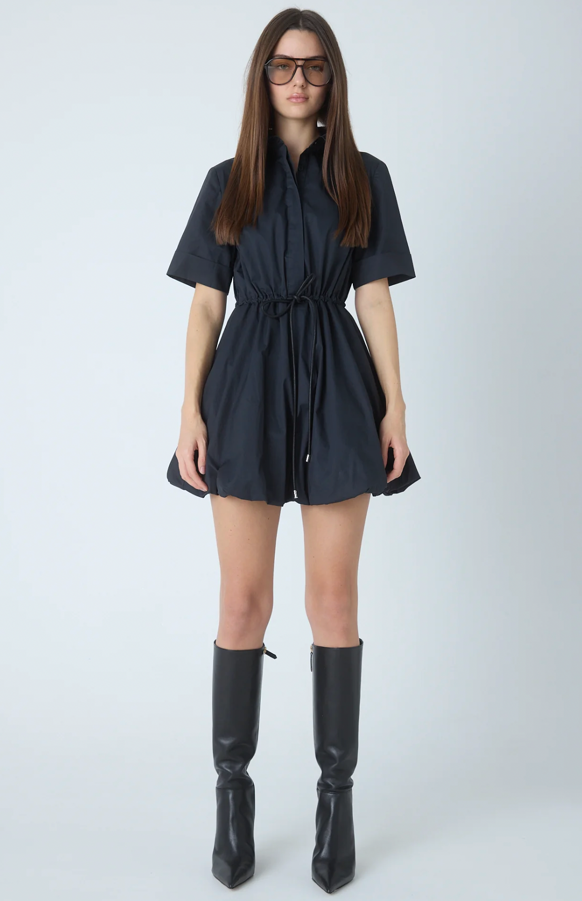 A woman modeling a short-sleeve black mini dress with a drawstring waist, paired with black knee-high boots, against a light gray studio background.