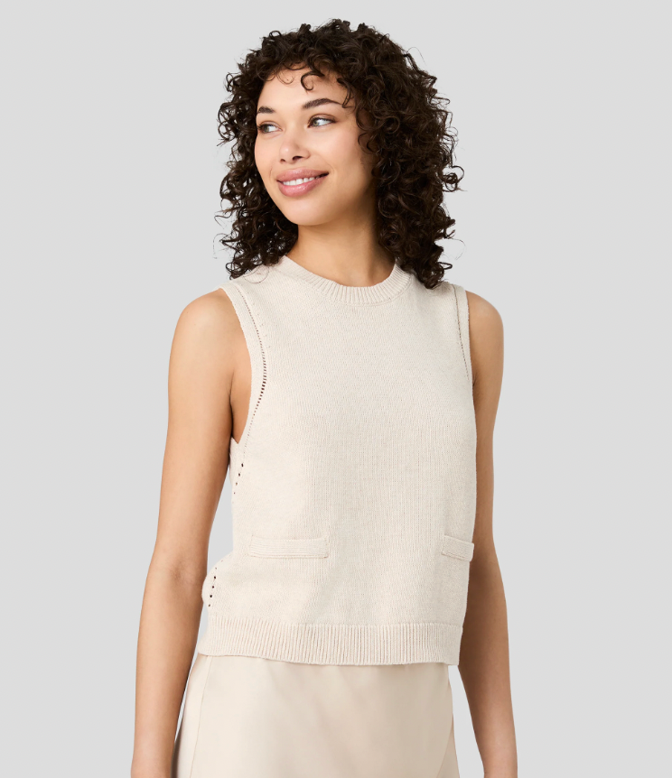 A woman with curly dark hair wearing a sleeveless cream knit vest with side cutouts and front pockets, paired with a matching cream skirt, against a solid light gray background.