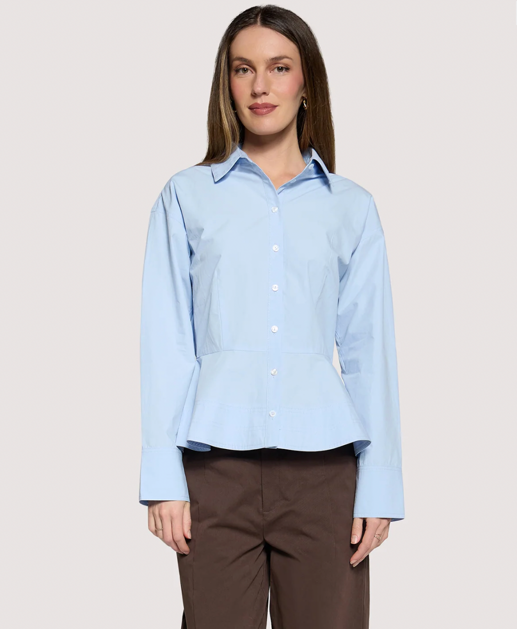 A woman modeling a light blue long-sleeve button-up blouse with a peplum waist, paired with brown trousers, against a solid light gray background.