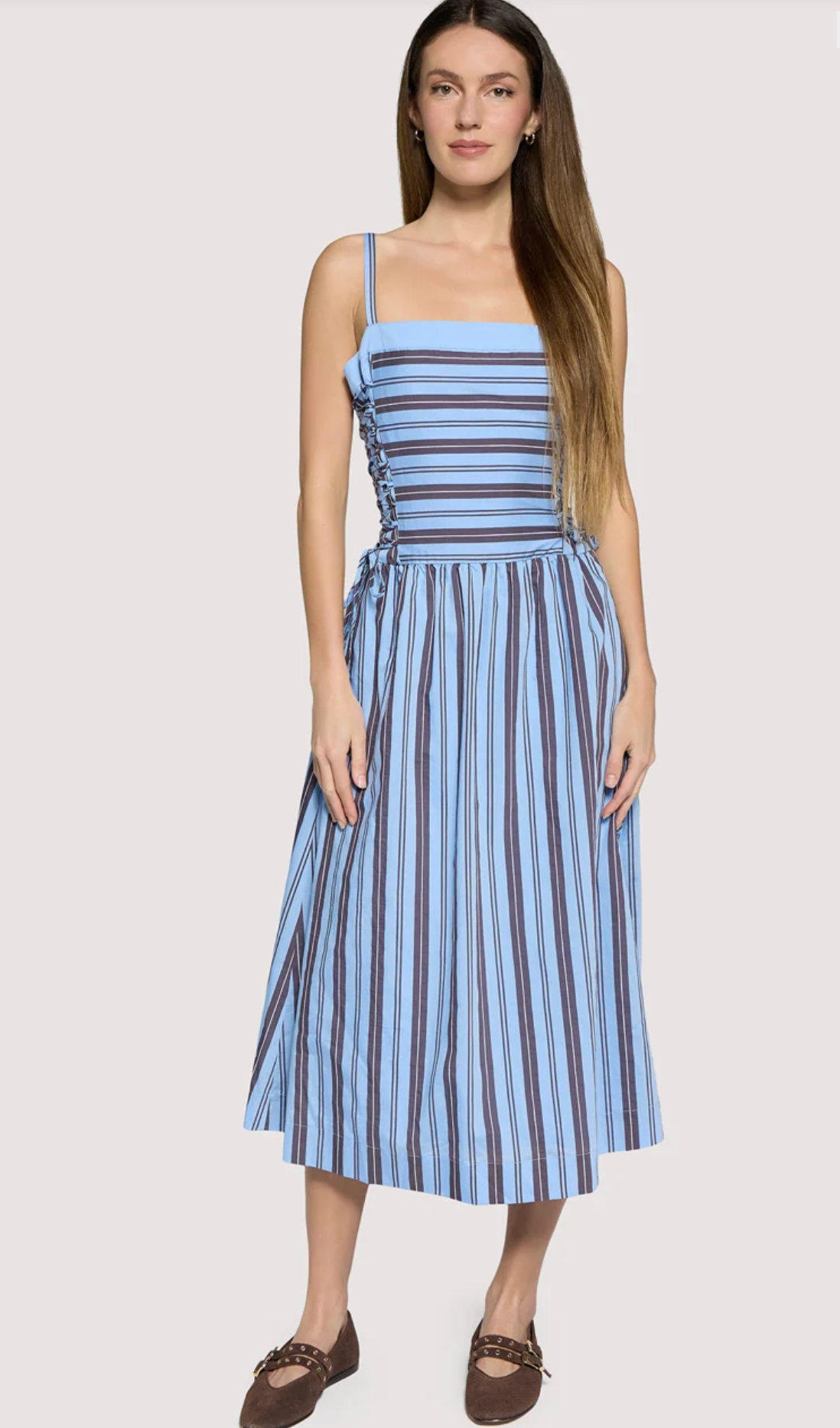 A woman modeling a sleeveless midi dress with thin shoulder straps, a gathered waist, and vertical blue, navy, and brown stripes on a light gray studio background.