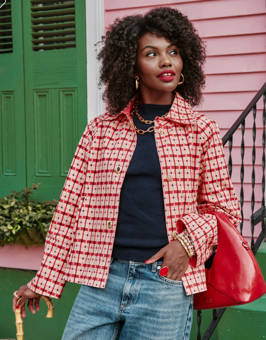 A woman stands outdoors in front of a pink house and green door, wearing a red-and-white patterned jacket over a navy top with blue jeans. She is holding a bright red handbag and a cane, with gold jewelry visible.