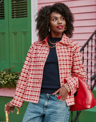 A woman stands outdoors in front of a pink house and green door, wearing a red-and-white patterned jacket over a navy top with blue jeans. She is holding a bright red handbag and a cane, with gold jewelry visible.
