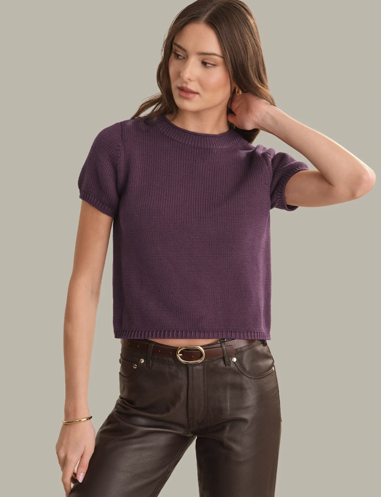 A woman modeling a short-sleeve ribbed knit sweater in a muted purple color, paired with dark brown pants, against a solid light gray background.