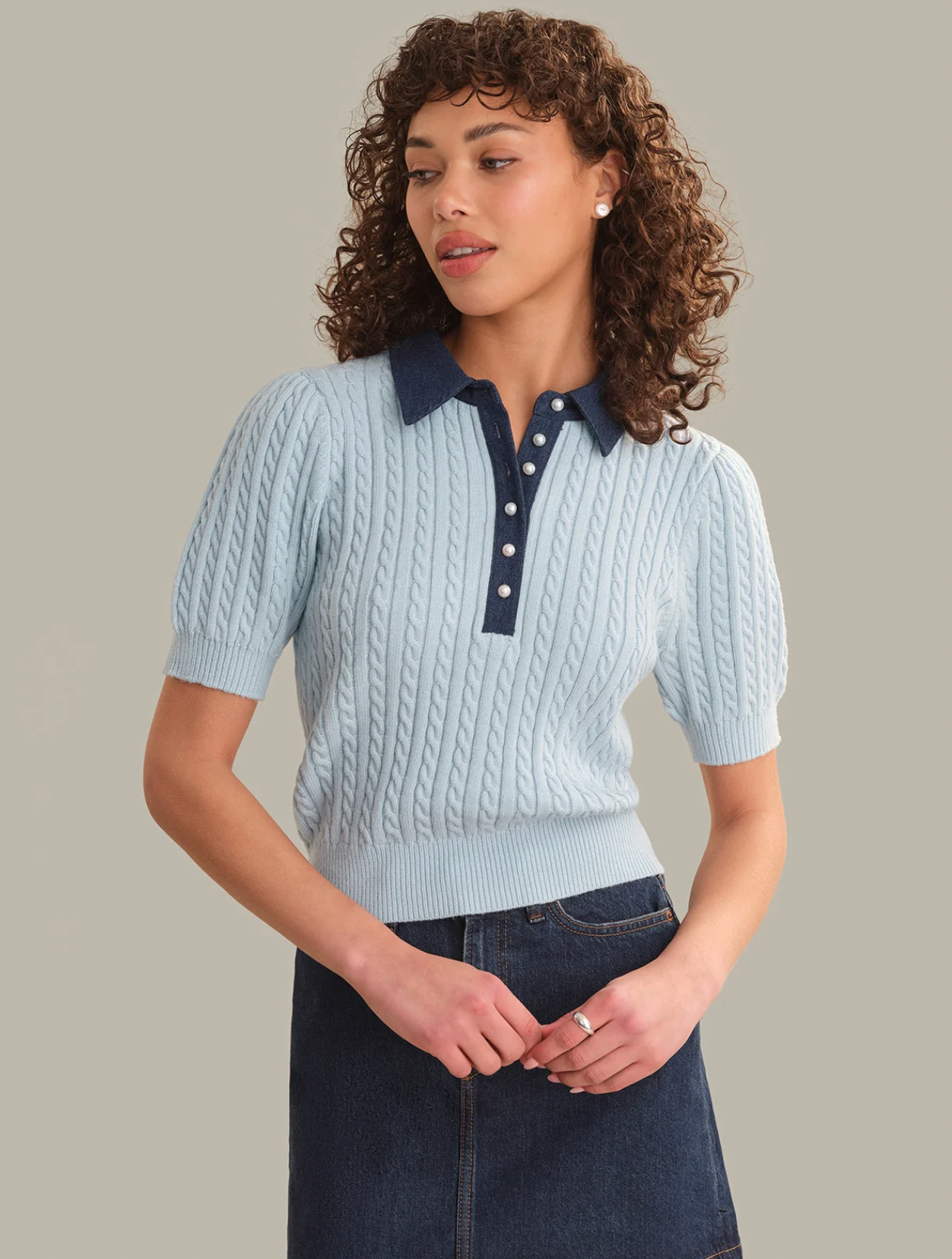 A woman modeling a light blue short-sleeve cable-knit polo sweater with a dark navy collar and button placket, paired with a dark denim skirt, against a solid beige background.