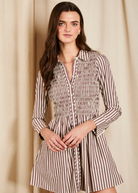 A woman modeling a brown and beige vertically striped mini dress with a smocked waist and long sleeves, standing against a neutral beige curtain backdrop.