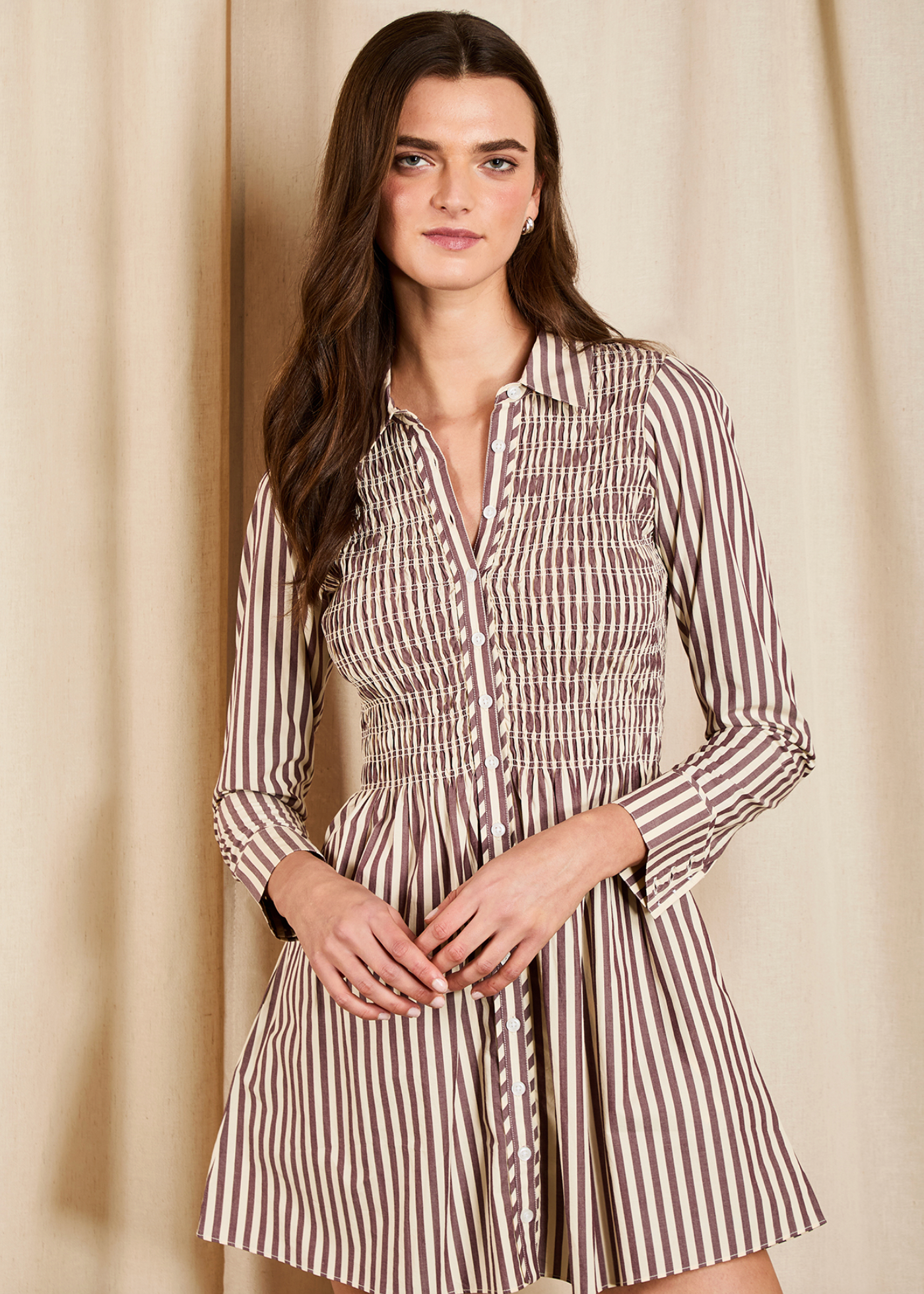 A woman modeling a brown and beige vertically striped mini dress with a smocked waist and long sleeves, standing against a neutral beige curtain backdrop.