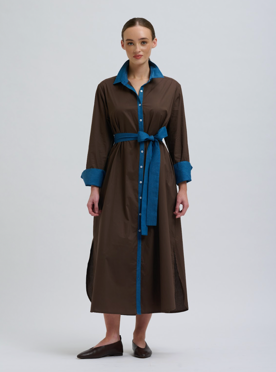 A woman modeling a brown long-sleeve midi shirt dress with a blue collar, cuffs, and waist tie belt, paired with dark brown leather shoes against a light gray studio background.