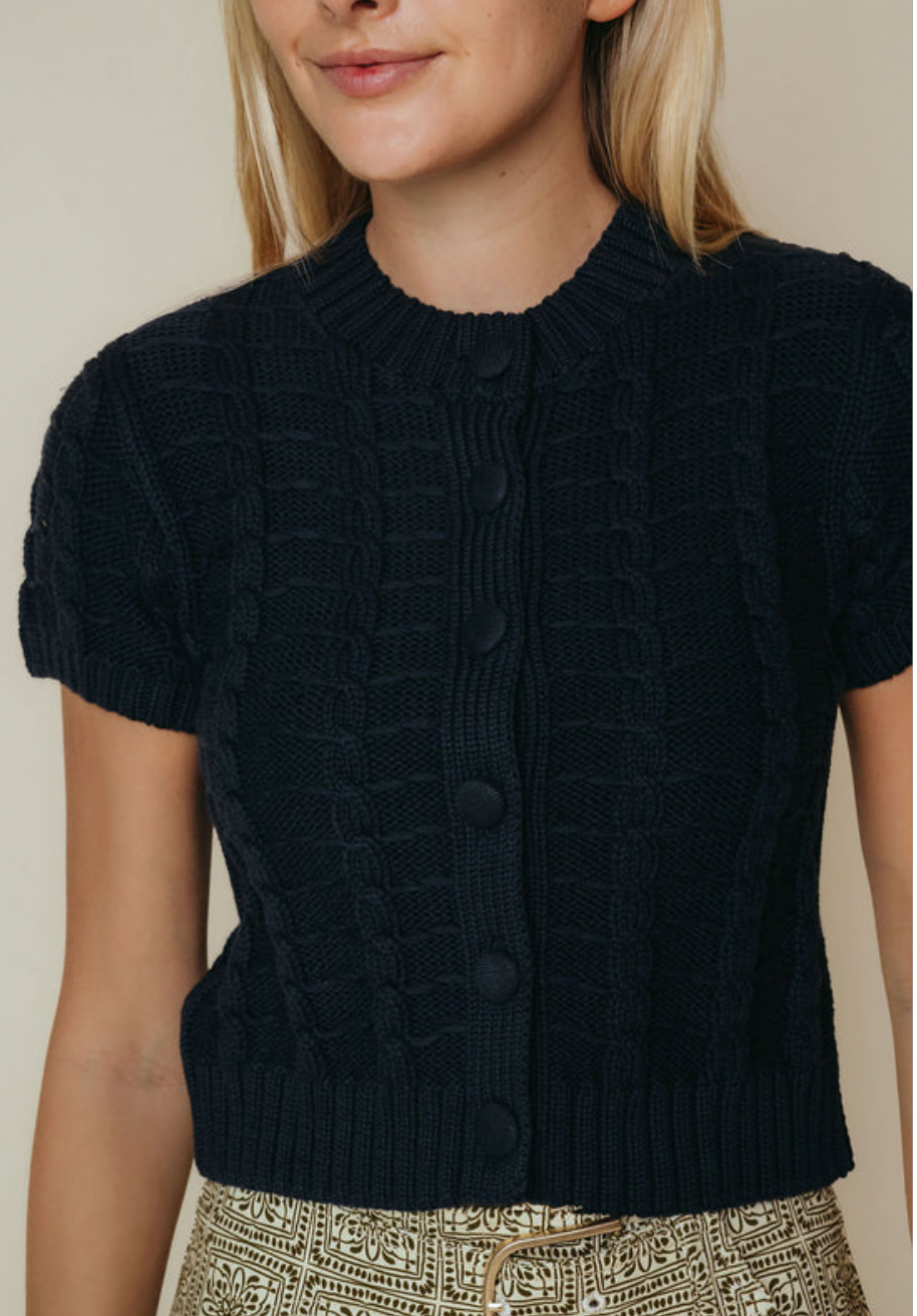 A woman wearing a dark navy blue short-sleeve cable-knit cardigan with a button-front closure, paired with patterned beige pants, against a plain beige background.