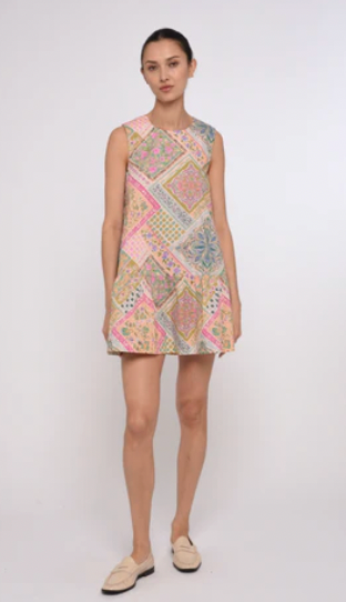 A woman modeling a sleeveless, knee-length A-line dress with a multicolor patchwork print in pink, beige, blue, and green, paired with beige loafers, against a plain light gray studio background.