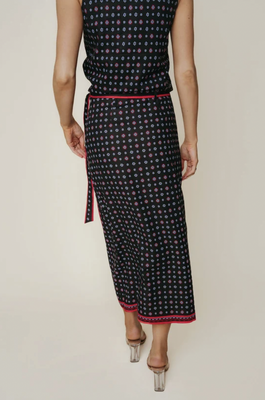 A model is shown from the back wearing a sleeveless black midi skirt with a colorful geometric pattern and a pink border, paired with clear high-heeled sandals against a light beige background.