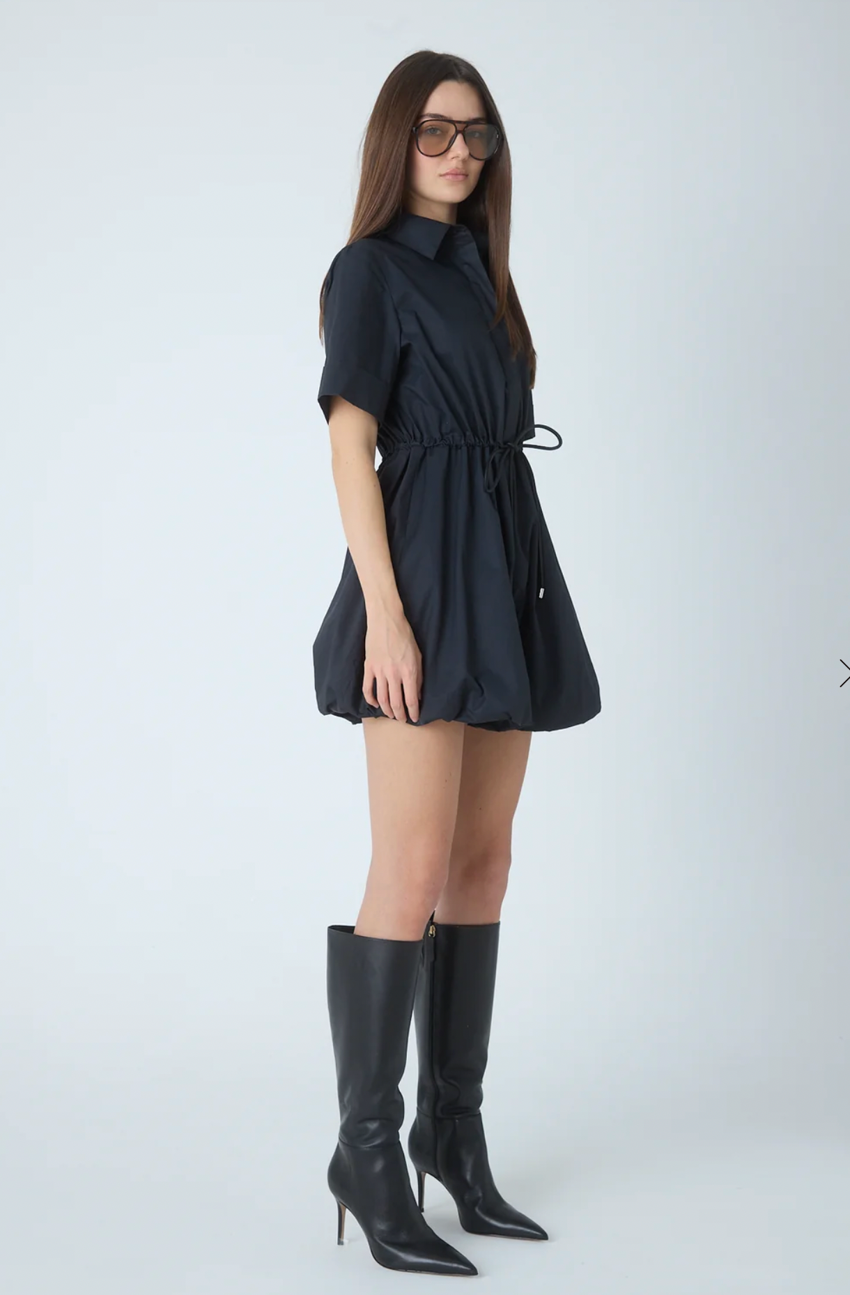 A woman modeling a short-sleeve black collared mini dress with a drawstring waist, paired with black knee-high heeled boots and sunglasses, against a light gray studio background.