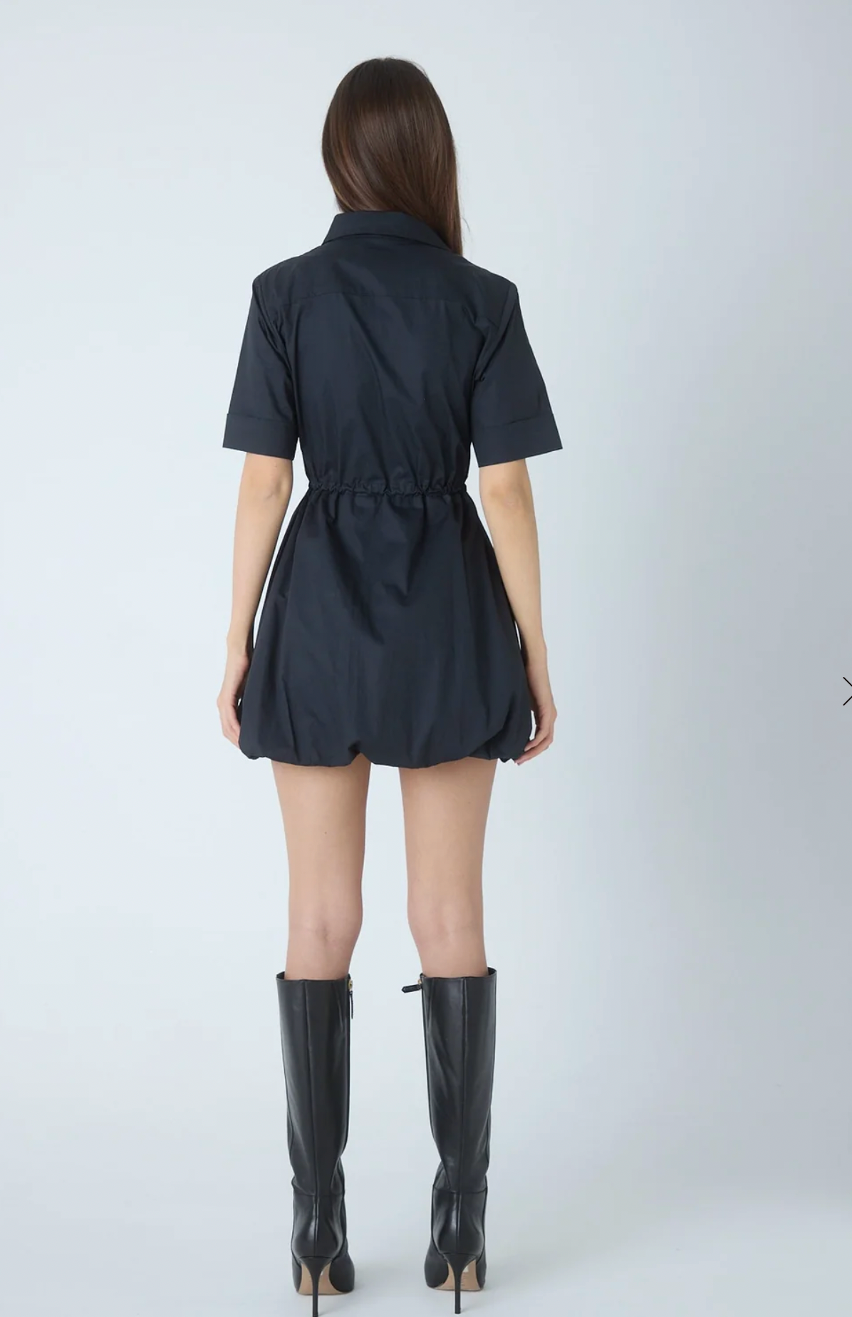 A woman shown from the back wearing a short-sleeve black mini dress with a tie waist and black knee-high heeled boots against a light gray studio background.