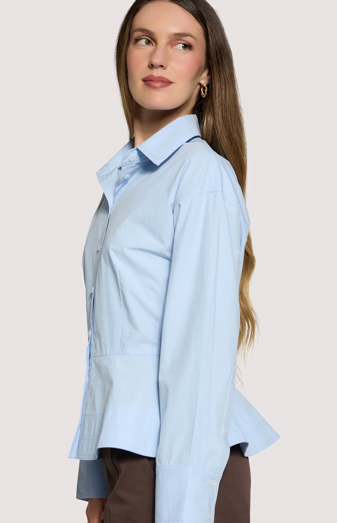 A woman wearing a light blue button-up shirt with a relaxed fit and peplum hem, shown in a side profile against a light beige studio background.