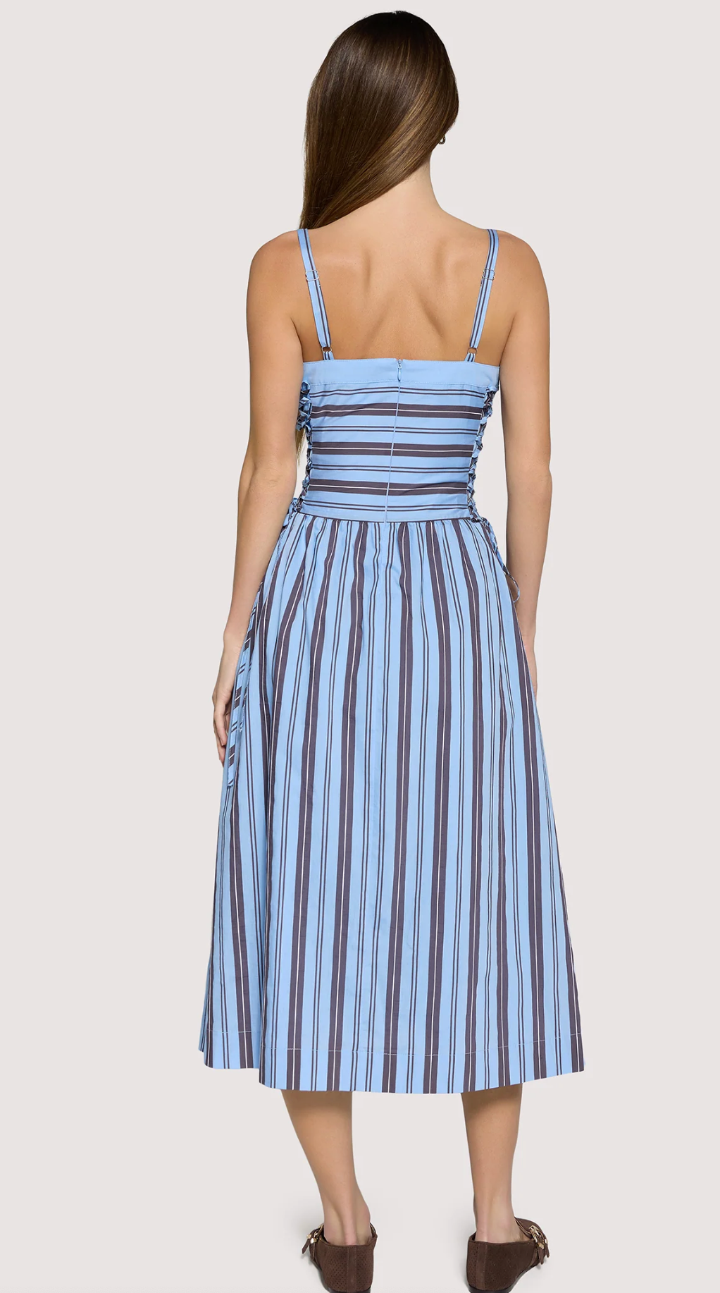 A woman shown from the back wearing a sleeveless midi dress with thin straps, a ruched bodice, and vertical blue, navy, and white stripes, photographed against a plain light gray studio background.