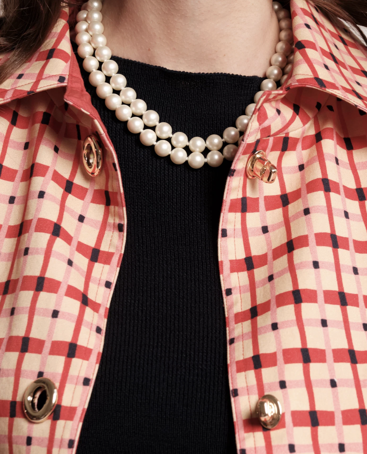Close-up of a person wearing a red and beige checkered button-up shirt over a black top, accessorized with a double-strand pearl necklace and gold-tone hardware.