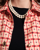 Close-up of a person wearing a red and beige checkered button-up shirt over a black top, accessorized with a double-strand pearl necklace and gold-tone hardware.