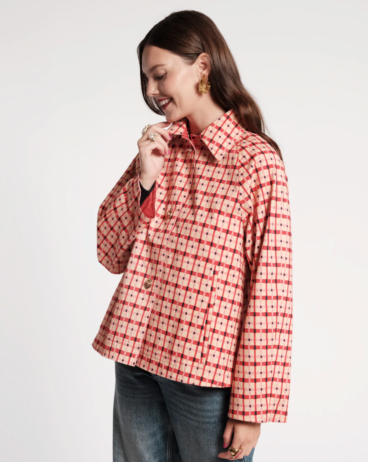 A woman wearing a red and white patterned button-up shirt with blue jeans, standing against a plain light gray studio background.