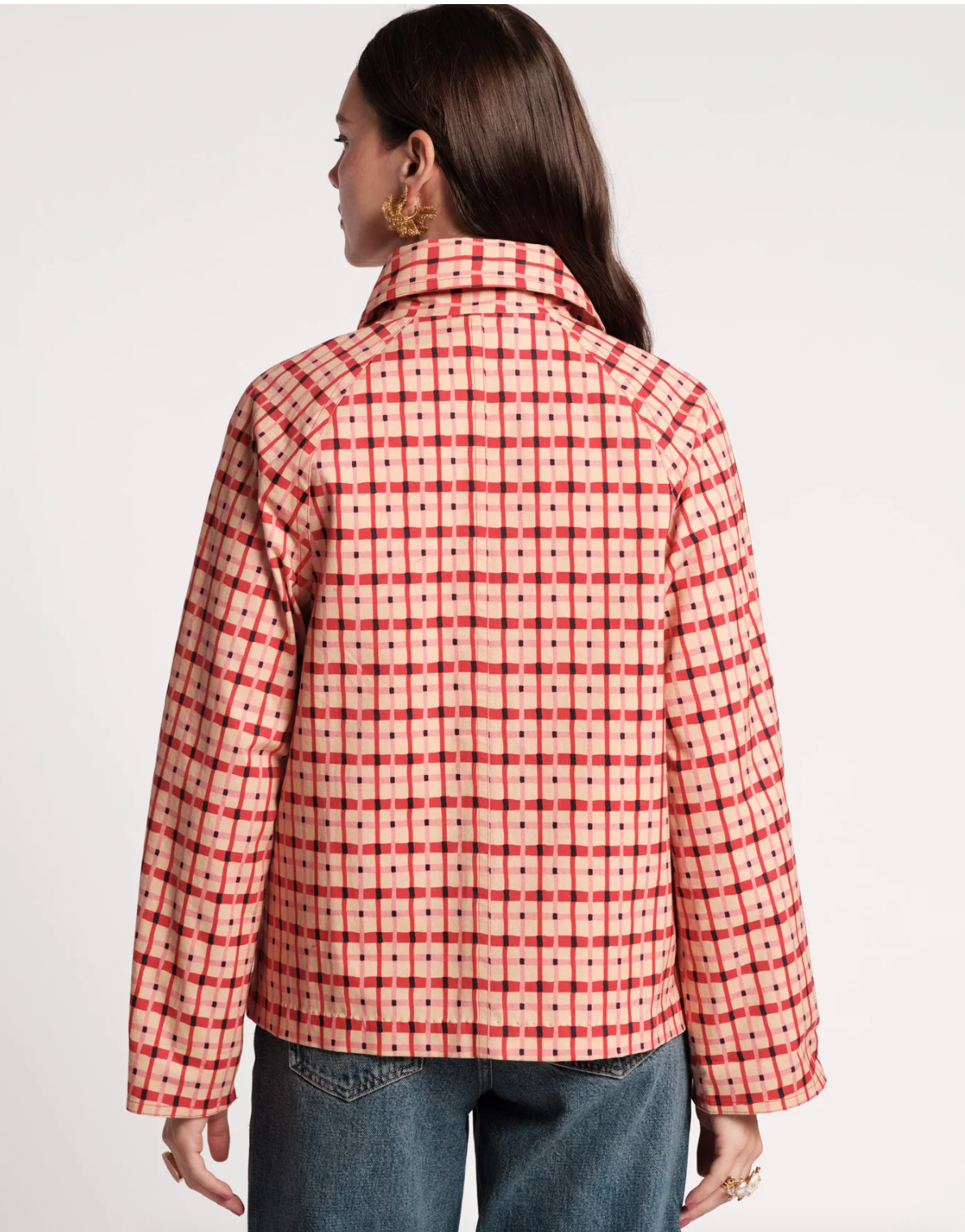A woman shown from the back wearing a red and white checkered patterned jacket with blue jeans, photographed against a plain light gray studio background.