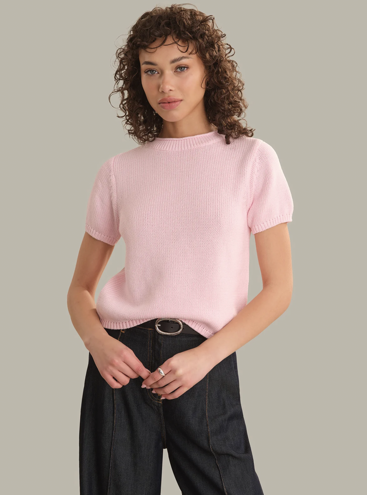 A woman with curly brown hair wearing a light pink short-sleeve ribbed knit sweater and dark wide-leg jeans, standing against a solid light gray background.