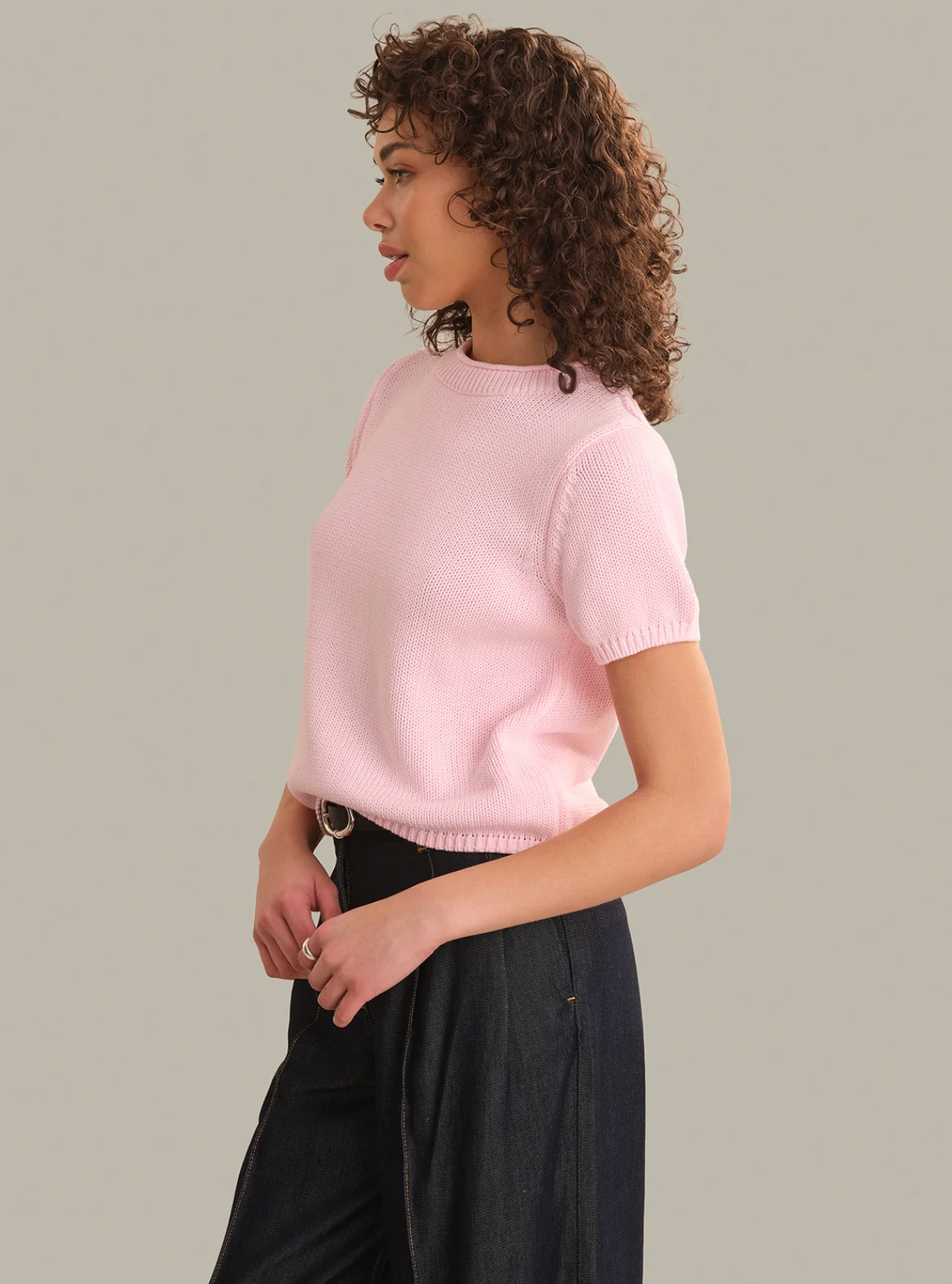 A woman with curly brown hair is shown in profile wearing a light pink short-sleeve knit top and dark wide-leg pants against a plain light gray studio background.