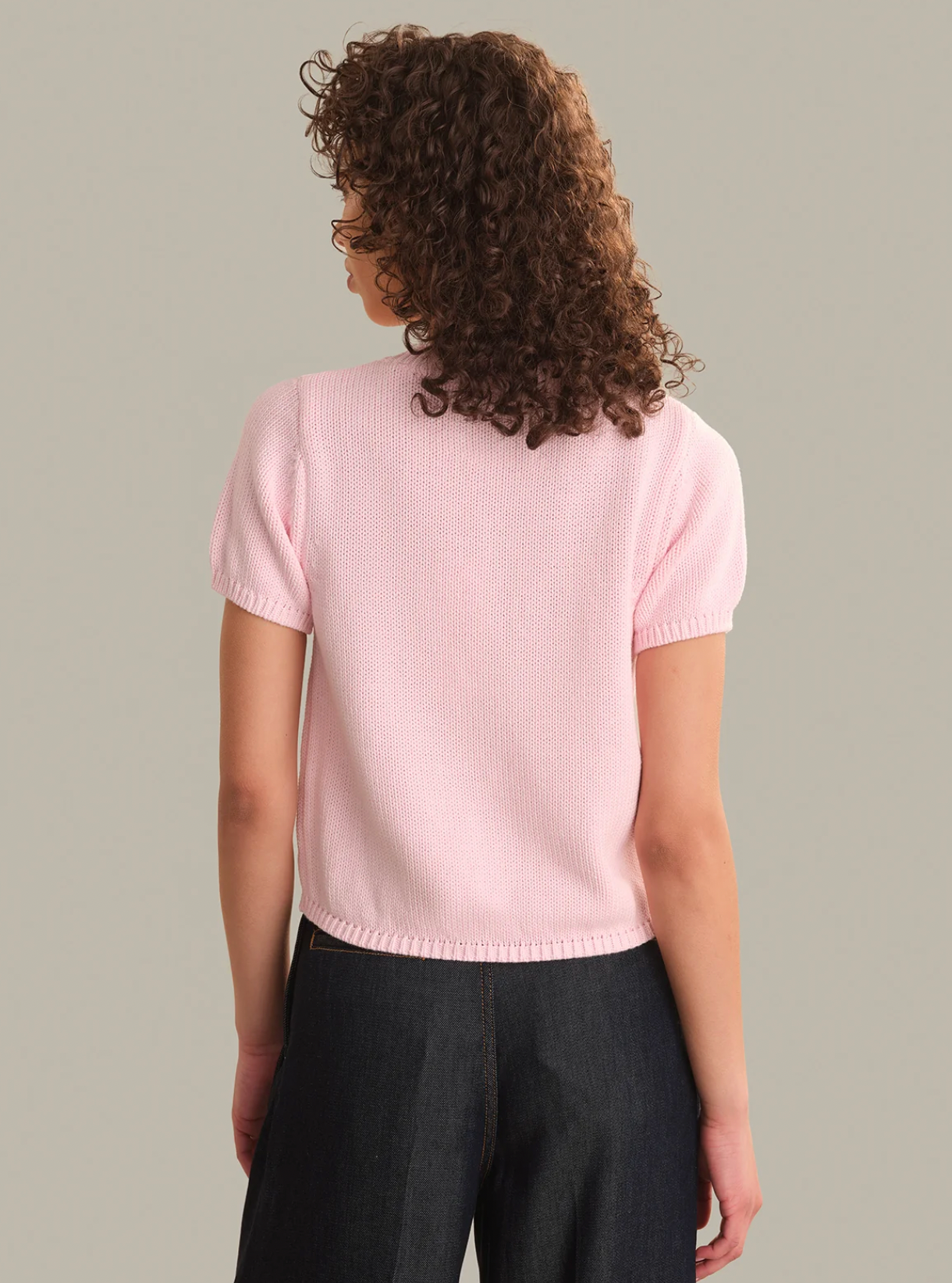 A woman with curly brown hair is shown from the back wearing a light pink short-sleeve knit top and dark wide-leg jeans against a solid light gray background.