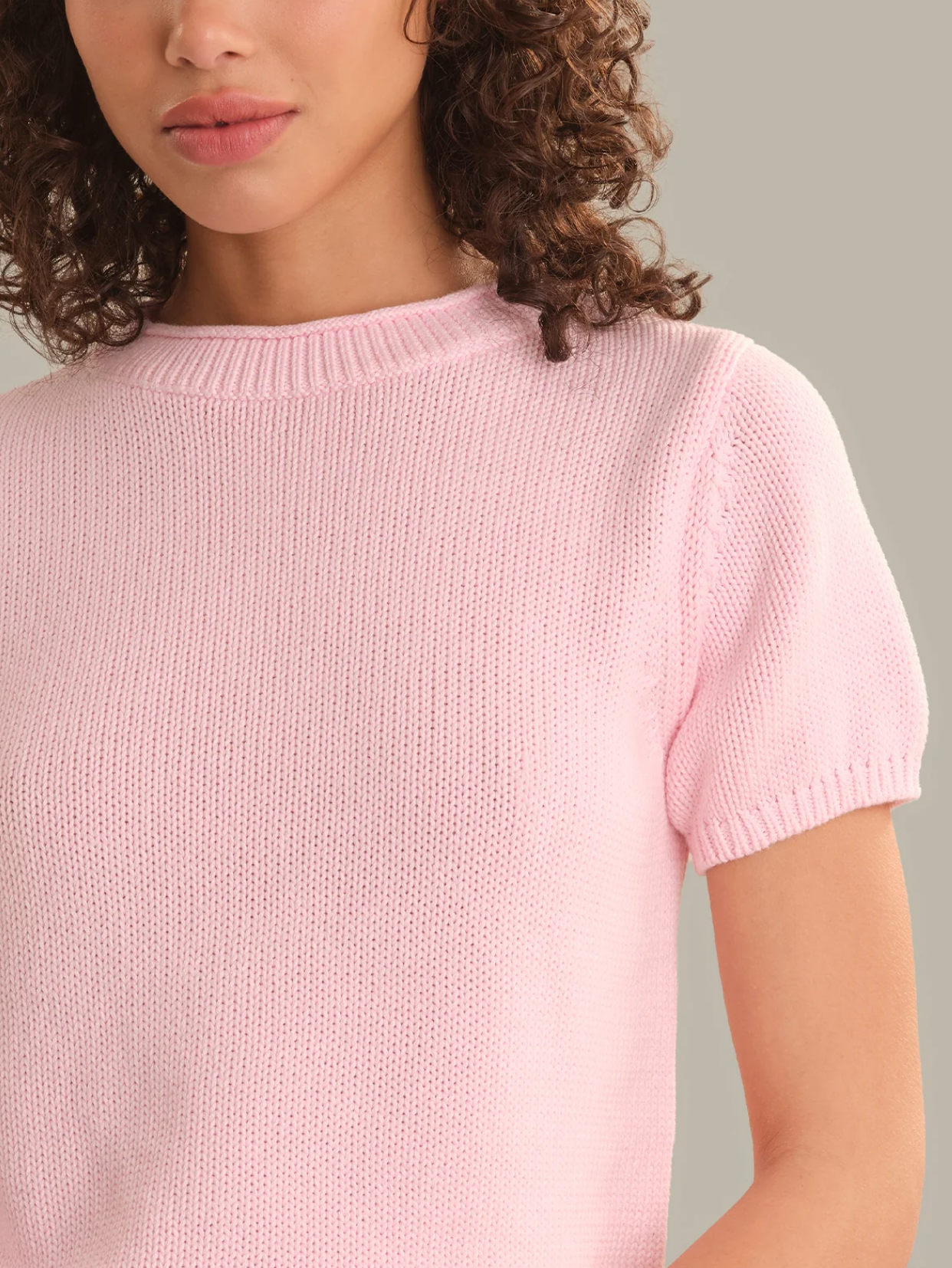 A close-up of a woman wearing a light pink short-sleeve knit top, showing the ribbed crew neck and textured knit fabric against a solid neutral background.