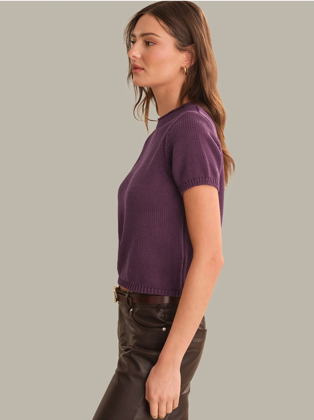 A woman shown in side profile wearing a short-sleeve ribbed knit top in purple and dark brown leather pants, against a solid light gray background.