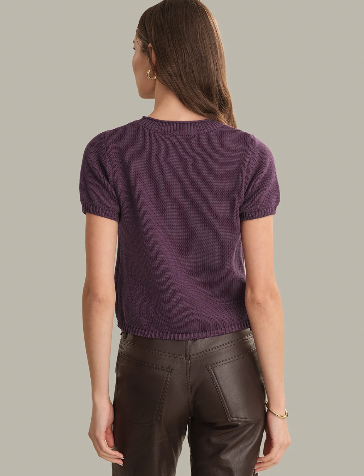 A woman shown from the back wearing a short-sleeve ribbed knit top in deep purple and dark brown faux-leather pants, set against a solid light gray background.