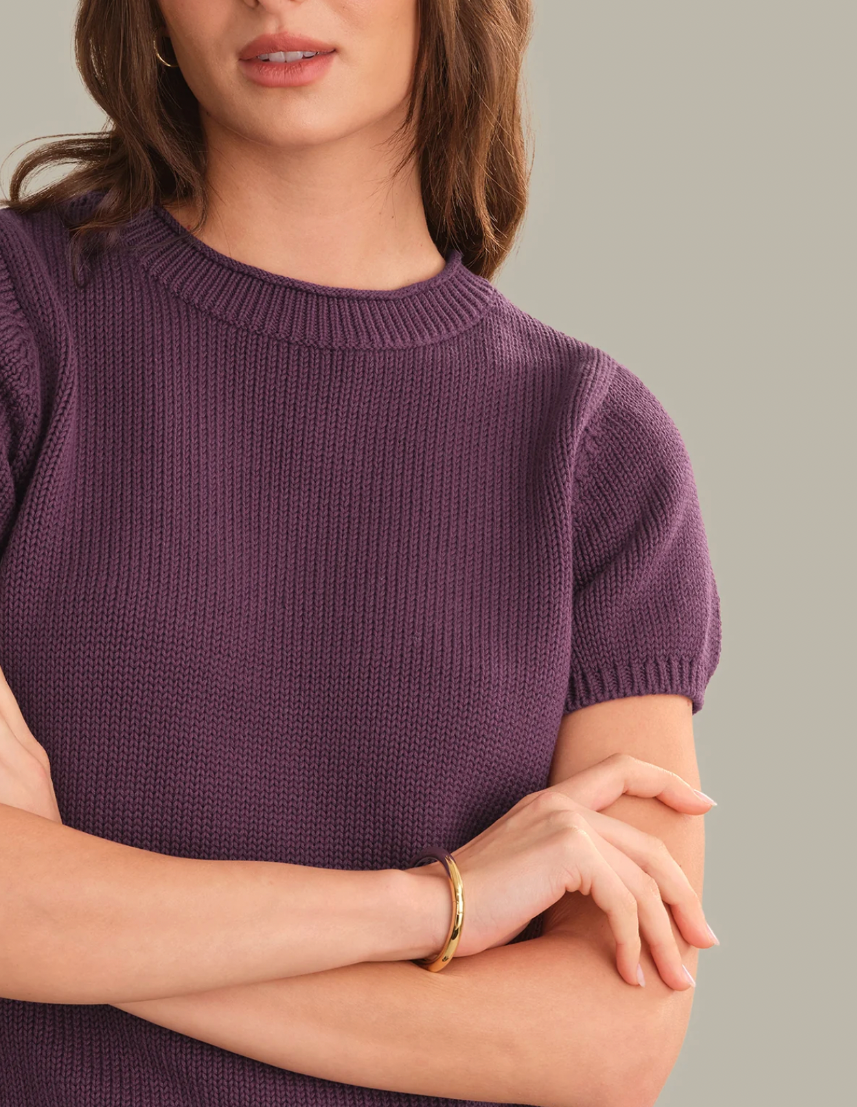 A close-up of a woman wearing a short-sleeve ribbed knit top in a muted purple color, with her arms crossed and a gold bangle bracelet visible on one wrist, against a solid light gray background.