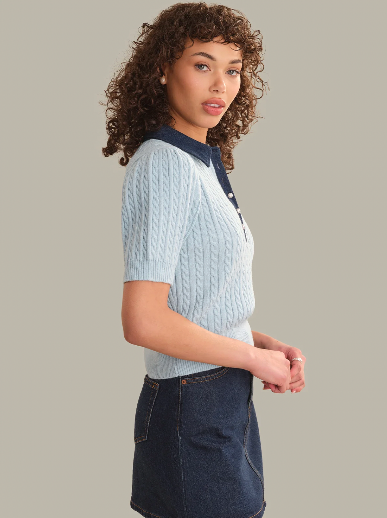 A woman modeling a light blue cable-knit short-sleeve polo sweater with a dark navy collar, paired with a dark denim skirt, against a solid light gray background.