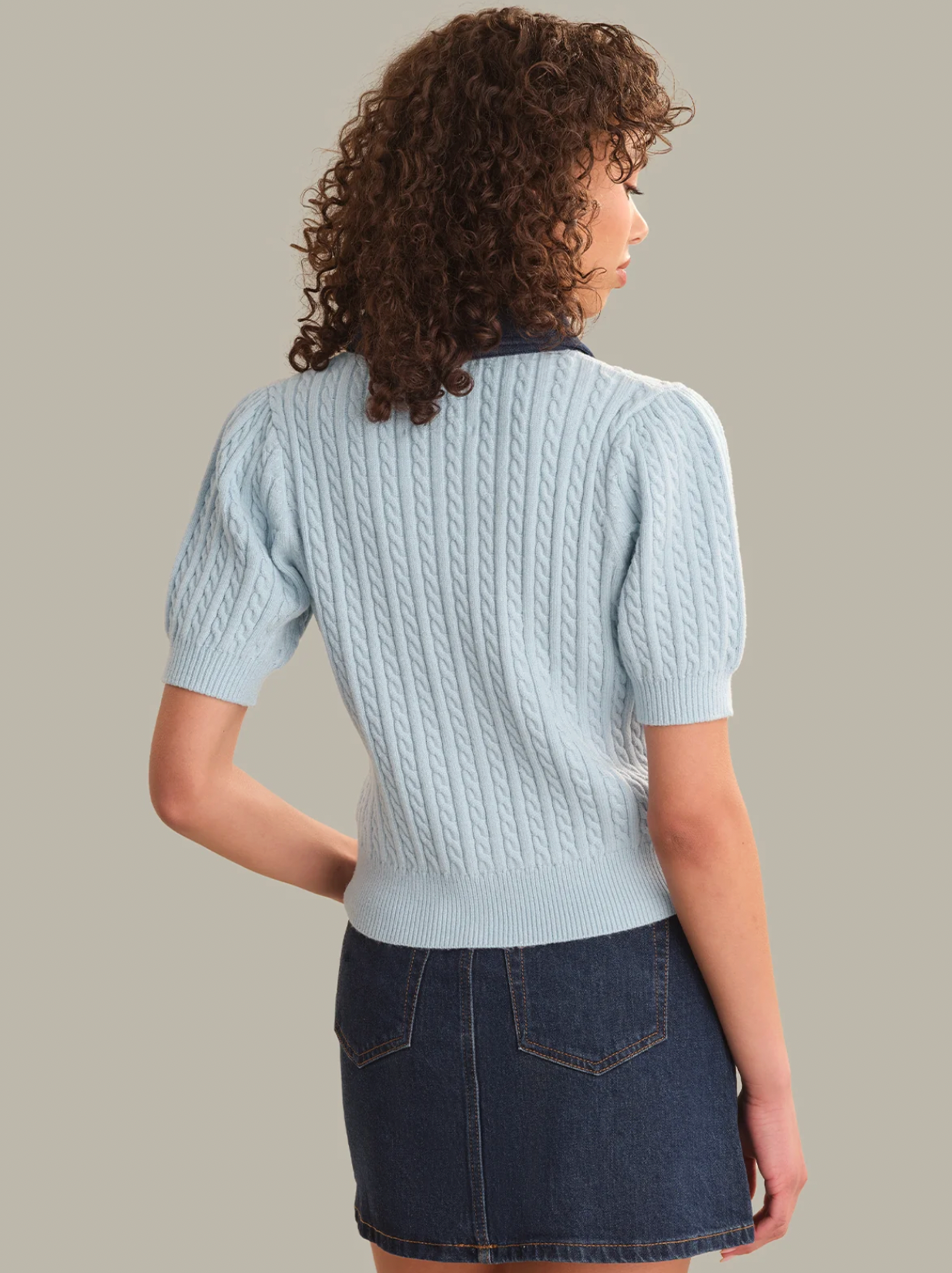 A model shown from the back wearing a light blue cable-knit short-sleeve sweater with a dark navy collar, paired with a dark denim mini skirt, against a solid beige background.