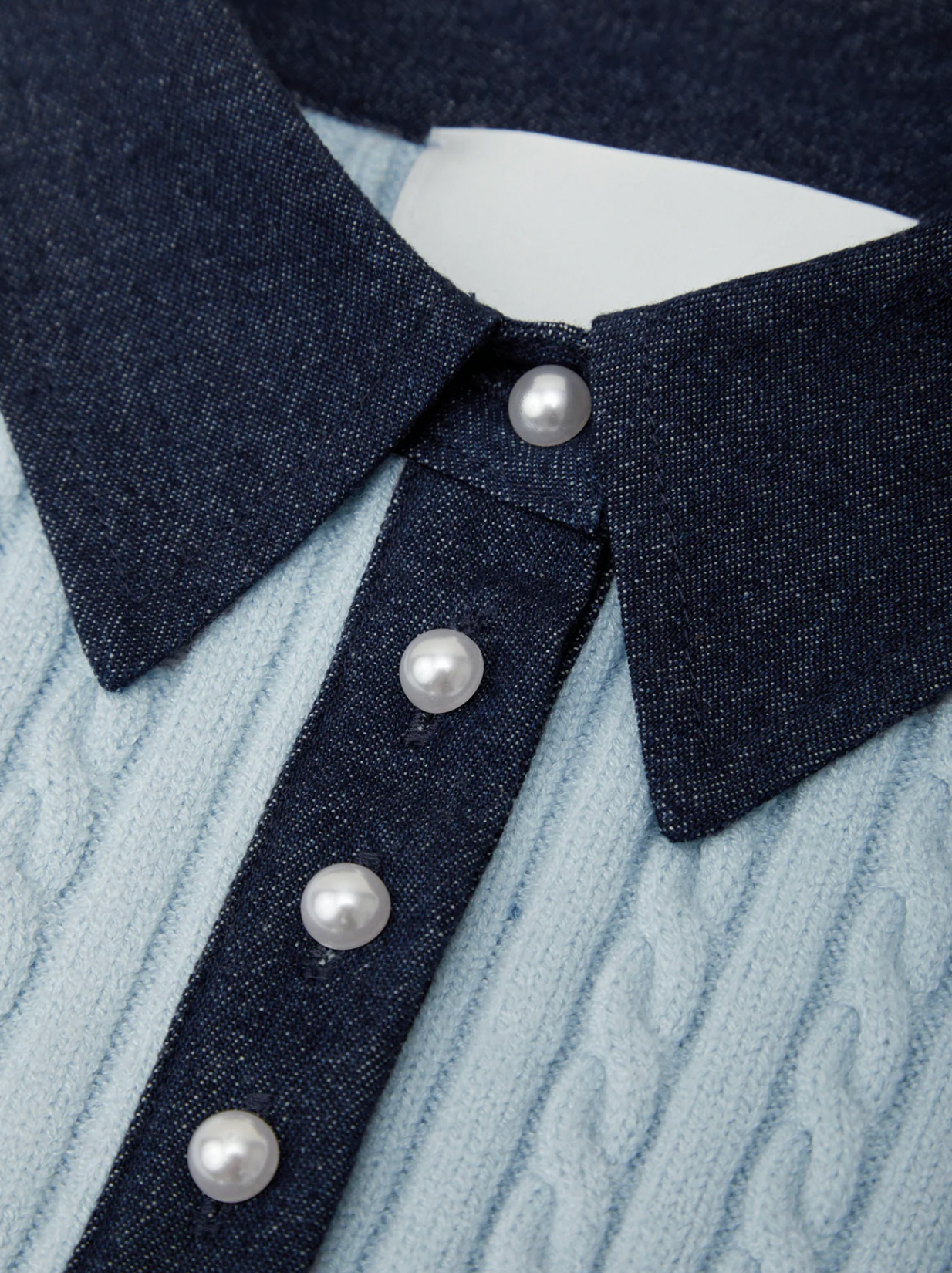 Close-up of a collared knit top with a dark navy denim-style collar and placket, decorated with four white pearl buttons, layered over a light blue cable-knit sweater.