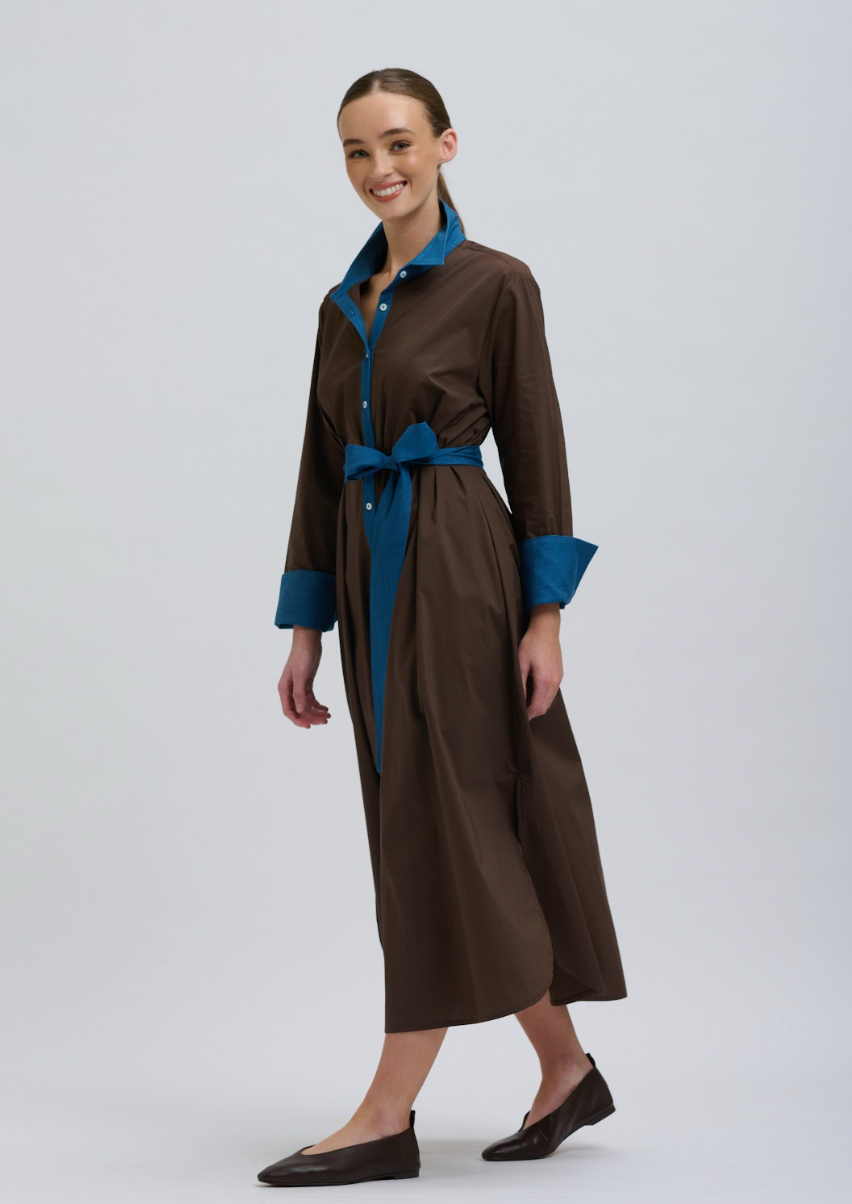 A woman modeling a brown long-sleeve midi shirt dress with a contrasting blue collar, cuffs, and waist tie, paired with dark brown flat shoes against a light gray studio background.