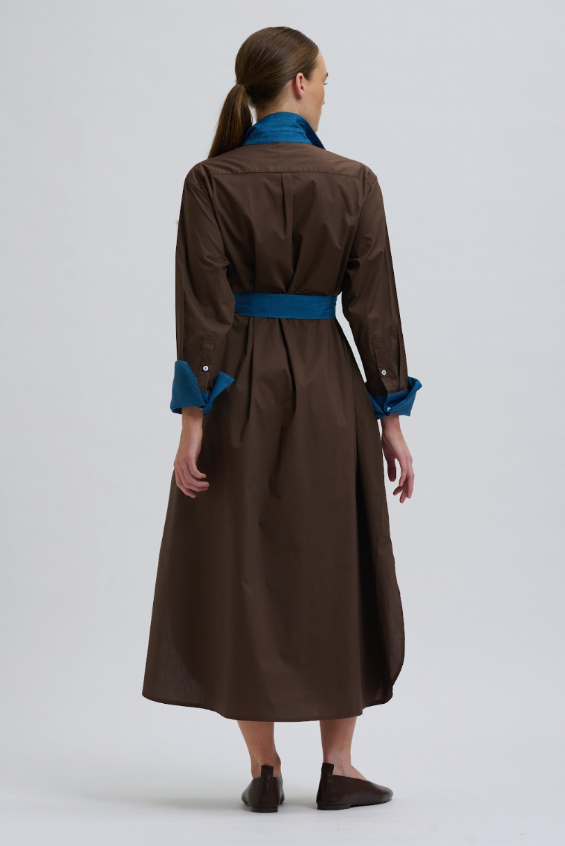 A woman shown from the back wearing a brown long-sleeve midi dress with a blue collar, blue waist belt, and blue turned-back cuffs, photographed against a light gray studio background.