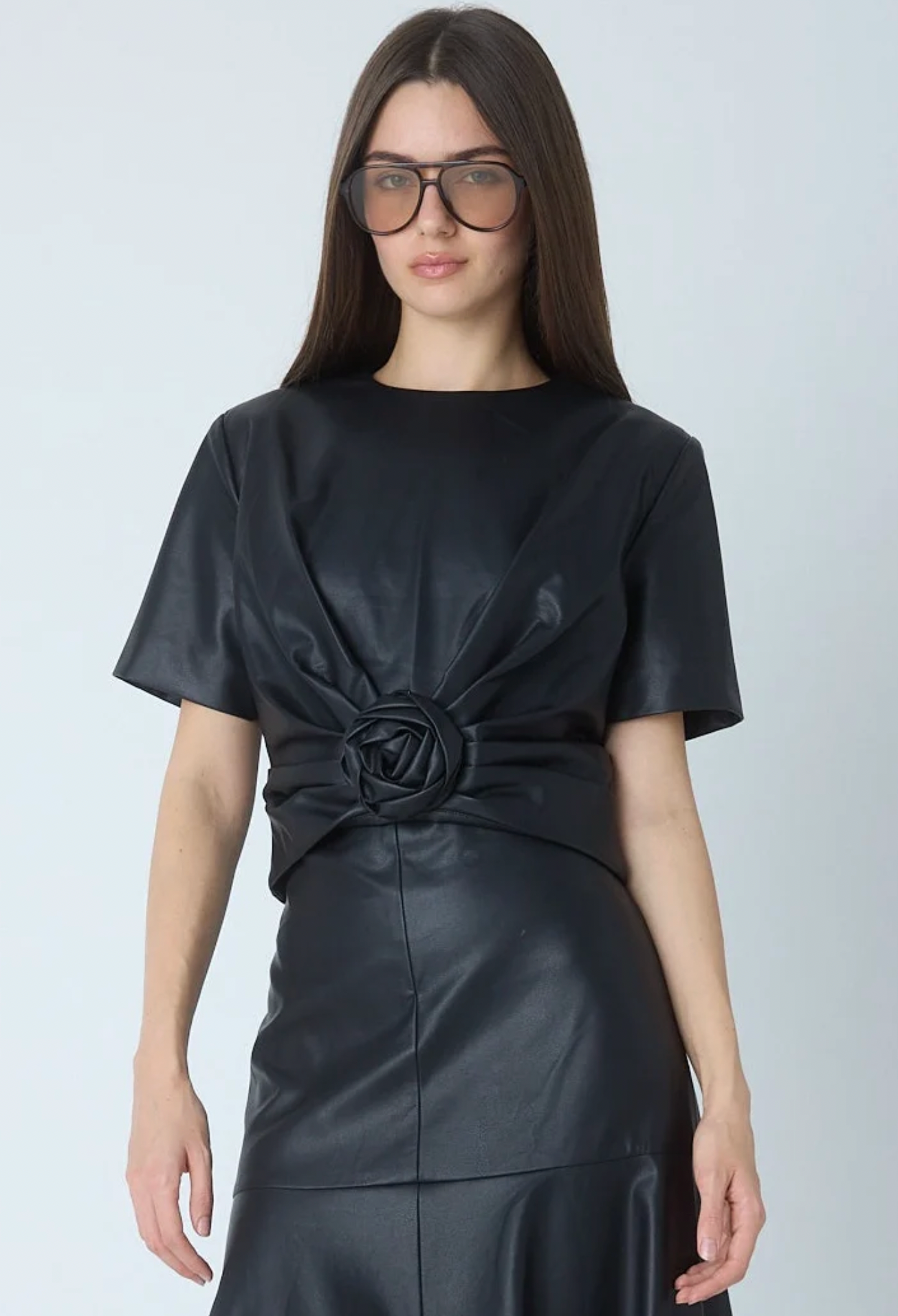A woman modeling a black short-sleeve dress with a gathered rose detail at the waist, paired with dark sunglasses, against a light gray studio background.