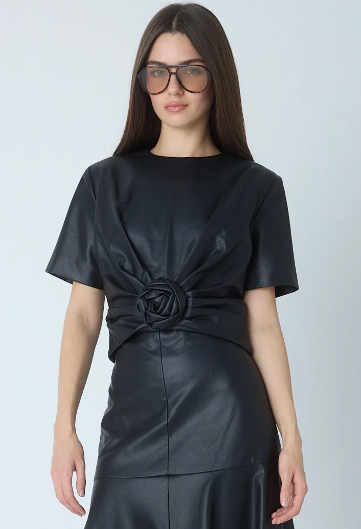 A woman modeling a black short-sleeve dress with a gathered rose detail at the waist, paired with dark sunglasses, against a light gray studio background.