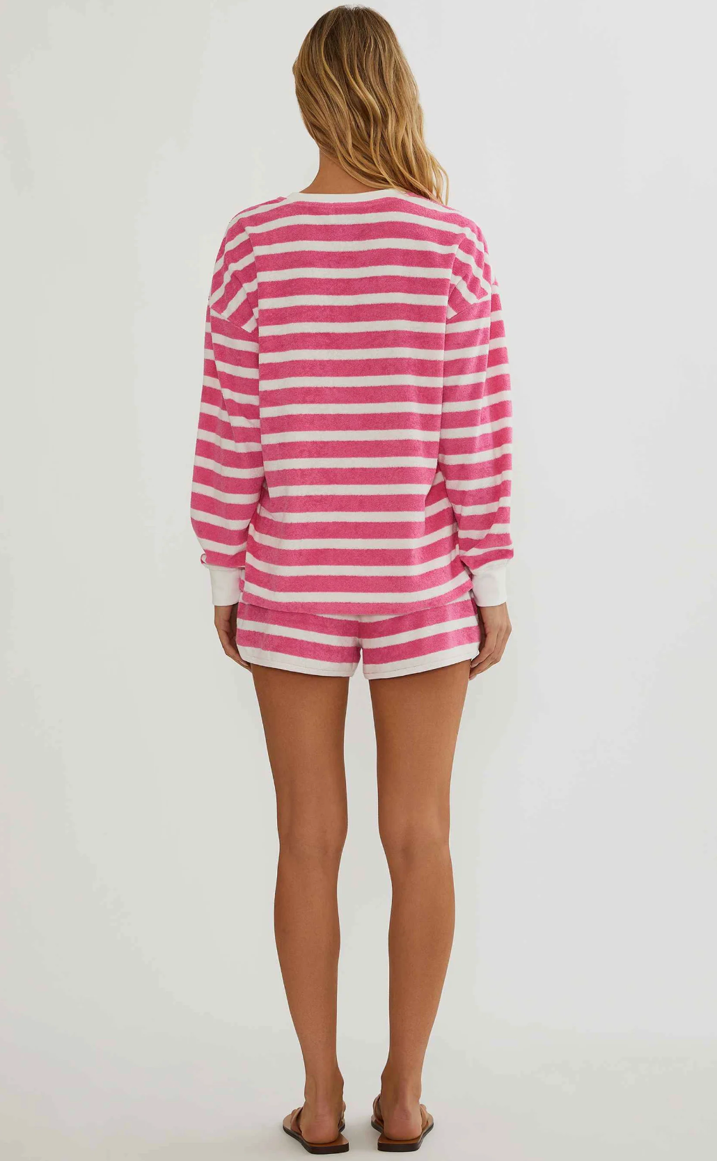 A woman viewed from behind wearing a pink and white horizontally striped long-sleeve top and matching shorts against a plain light gray studio background.