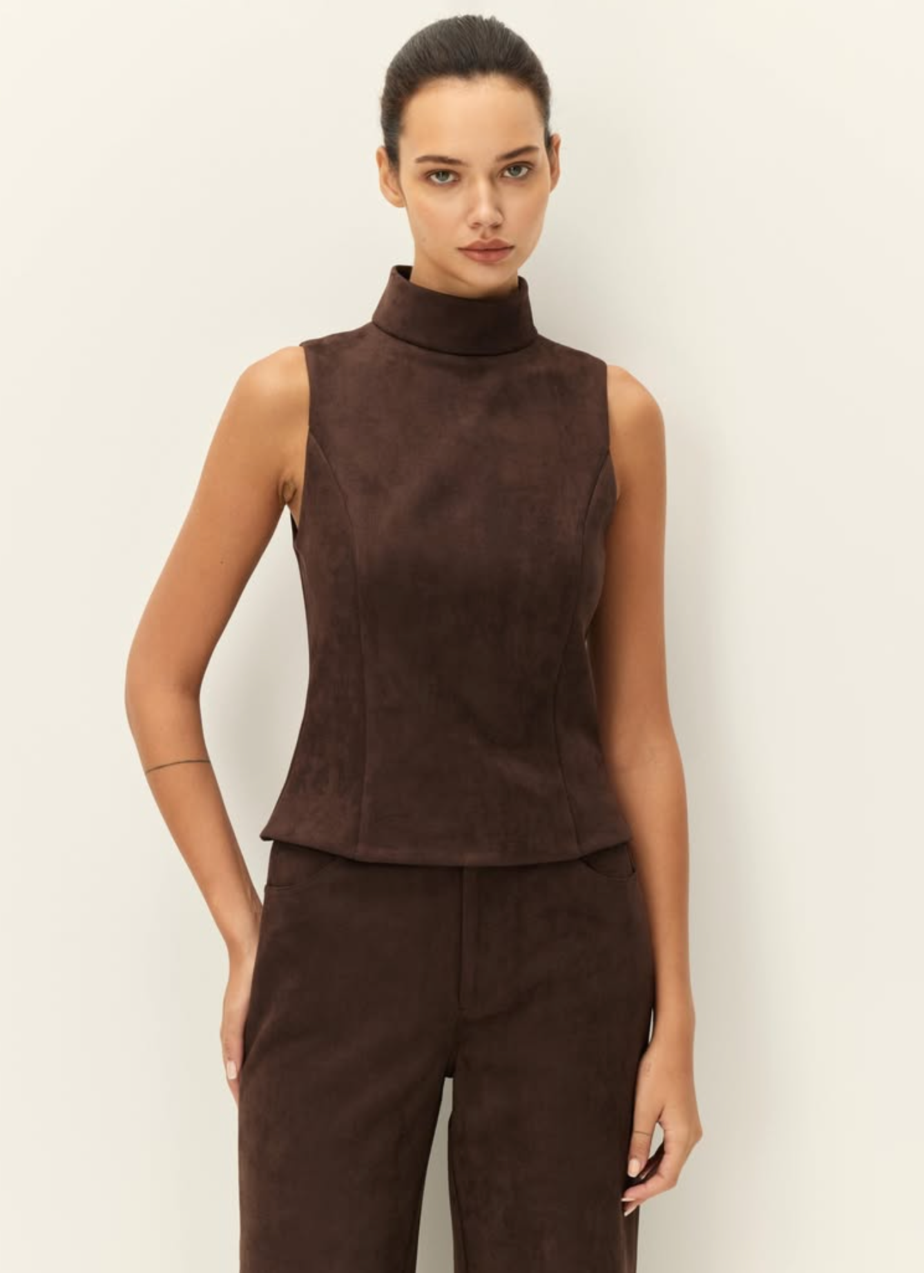 A model wearing a sleeveless, high-neck brown faux-suede top and matching brown trousers, standing against a plain off-white background.