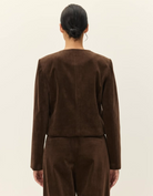 A model shown from the back wearing a dark brown cropped jacket and matching trousers, photographed against a plain off-white studio background.