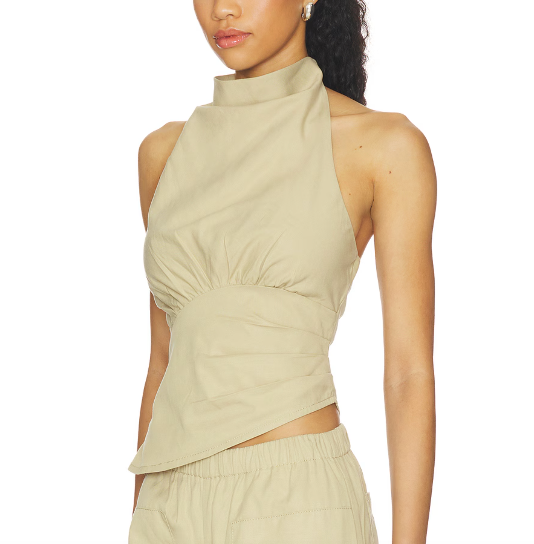 A model wearing a beige halter crop top with gathered detailing and a high mock neck, paired with matching beige pants against a white background.