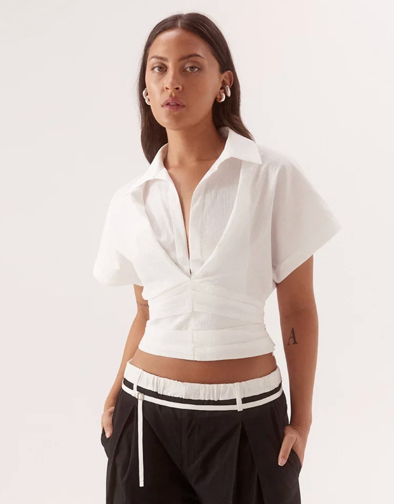 A woman modeling a white short-sleeve cropped button-up shirt with a twisted front detail, paired with black pants featuring a white stripe, against a plain white studio background.